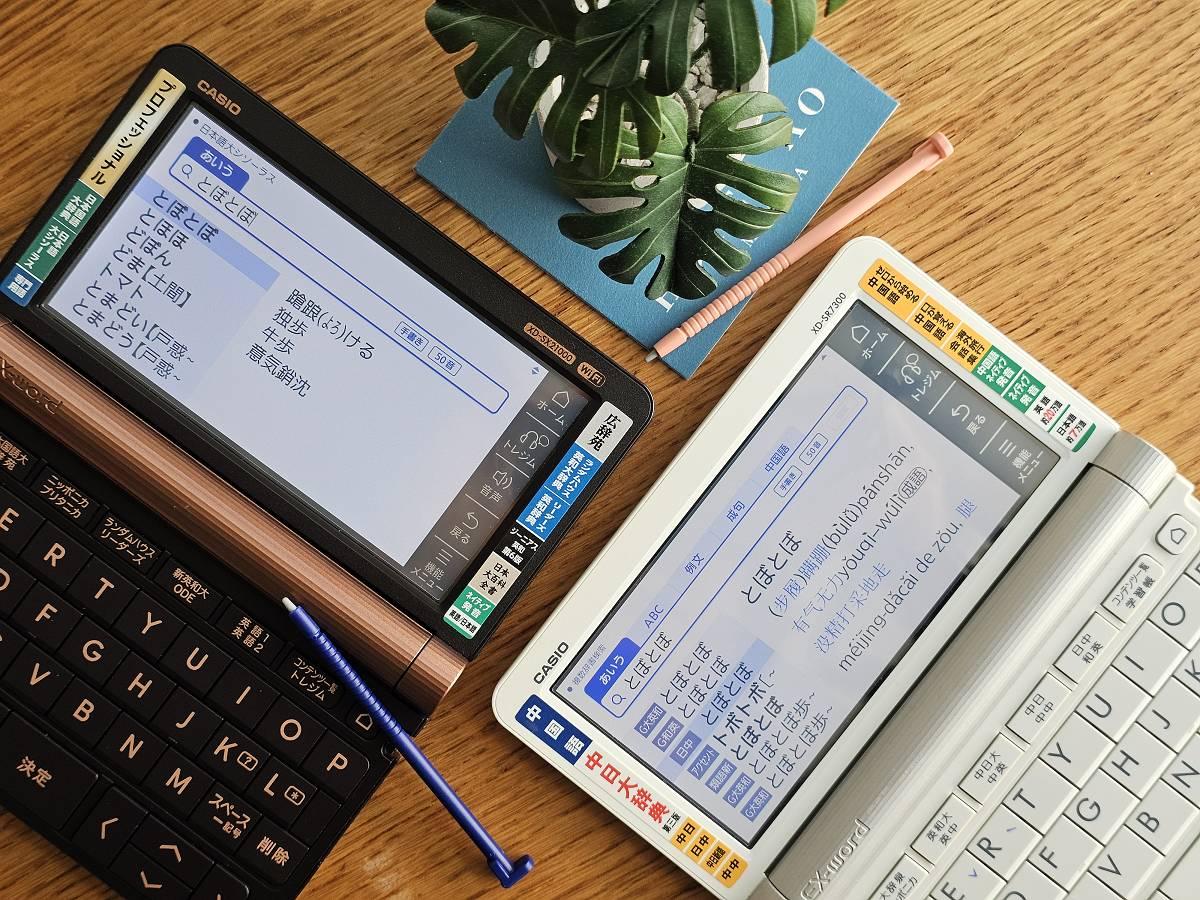 木質桌面上並排兩台 CASIO 電子辭典（左為深色機身 XD-SX21000，右為白色機身 XD-SR7300），螢幕分別顯示日文同義詞查詢結果與中日辭典翻譯內容，一旁擺著多肉植物與筆記本。