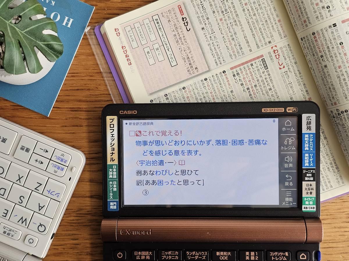 CASIO XD-SX21000 電子辭典螢幕顯示古語辭典「わびし」一詞的例句與白話翻譯，一旁攤開的紙本辭典也翻到同一詞條的頁面。