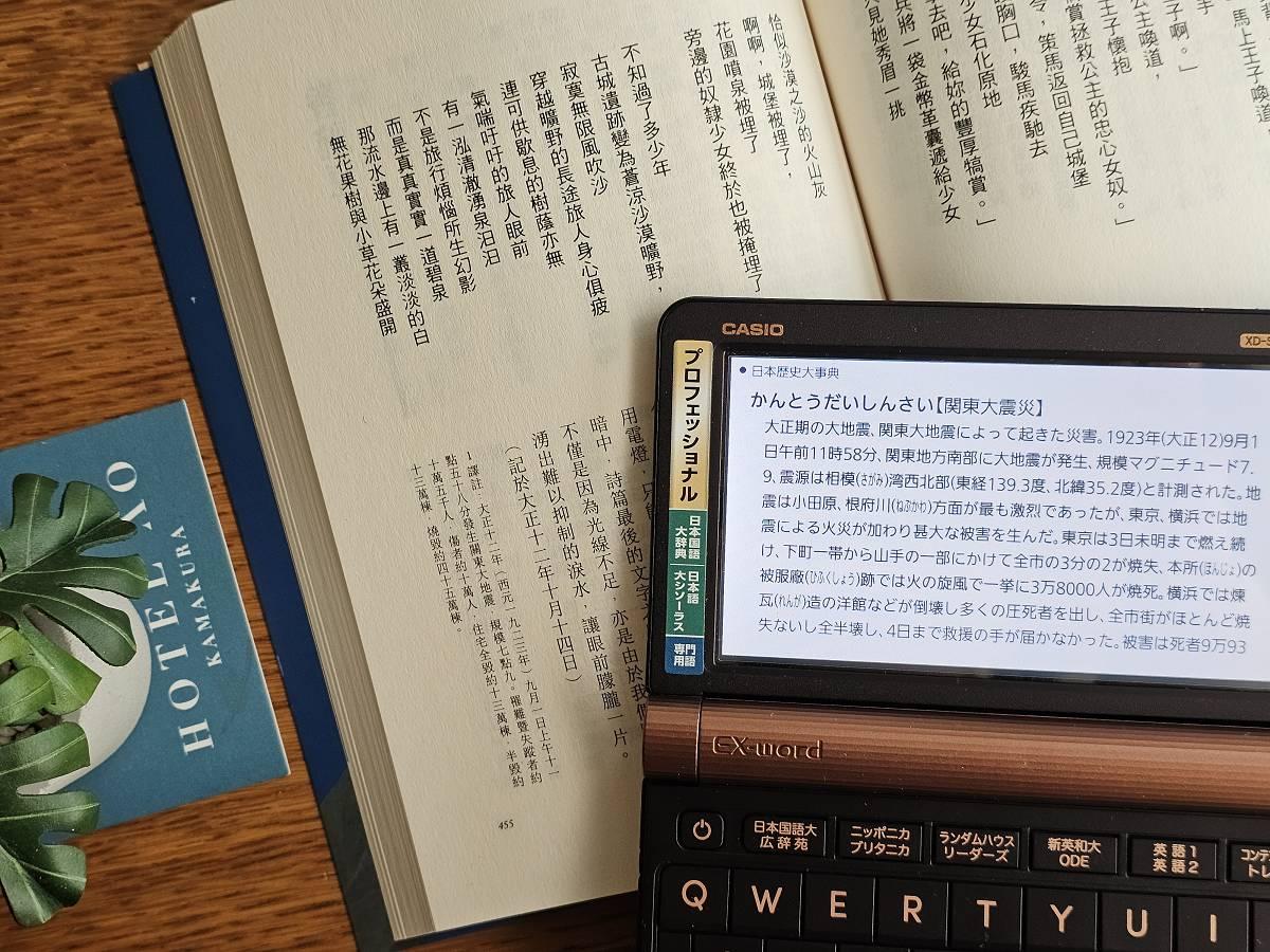 CASIO 電子辭典螢幕顯示「關東大震災」的百科辭典解說，機身斜放在翻開的中文譯本小說書頁上，一旁露出飯店資料夾與多肉植物。