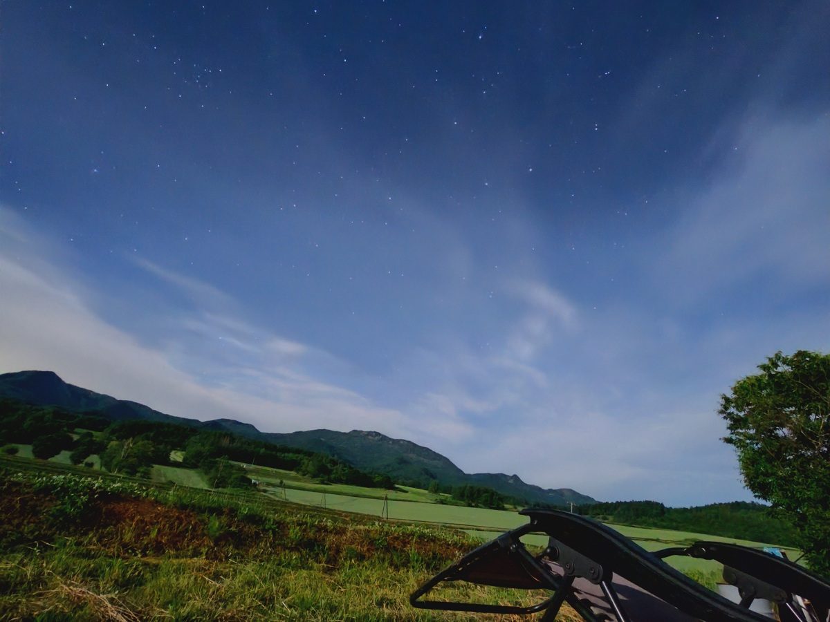 富良野夜空繁星點點,薄雲飄過深藍色天幕,遠方是連綿山脈與大片農田,前景可見一張戶外躺椅的椅腳與一旁的樹影。