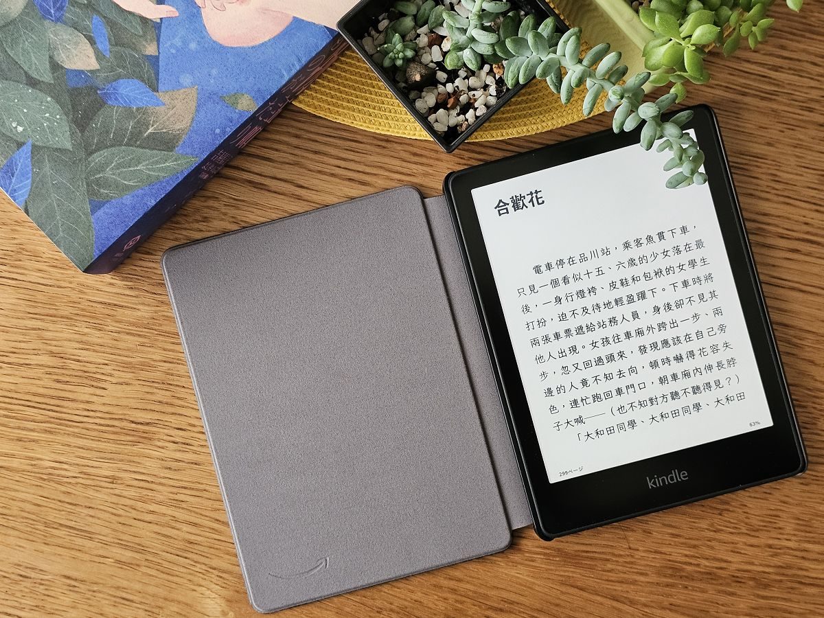 攤開灰色保護殼的 Kindle Paperwhite Kids 立在木桌上，螢幕顯示吉屋信子《花物語》繁體中文版〈合歡花〉篇章內文，周圍擺著多肉植物與一本插畫書籍。