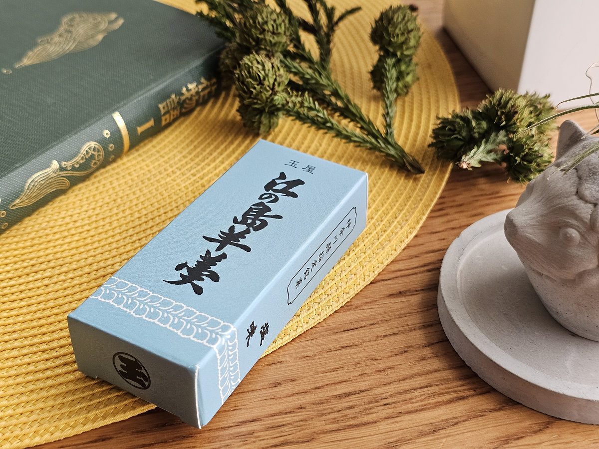 淺藍色包裝的「玉屋 江之島羊羹」紙盒橫放在黃色編織桌墊上，一旁有精裝日文書籍、乾燥花束與陶製小擺飾。